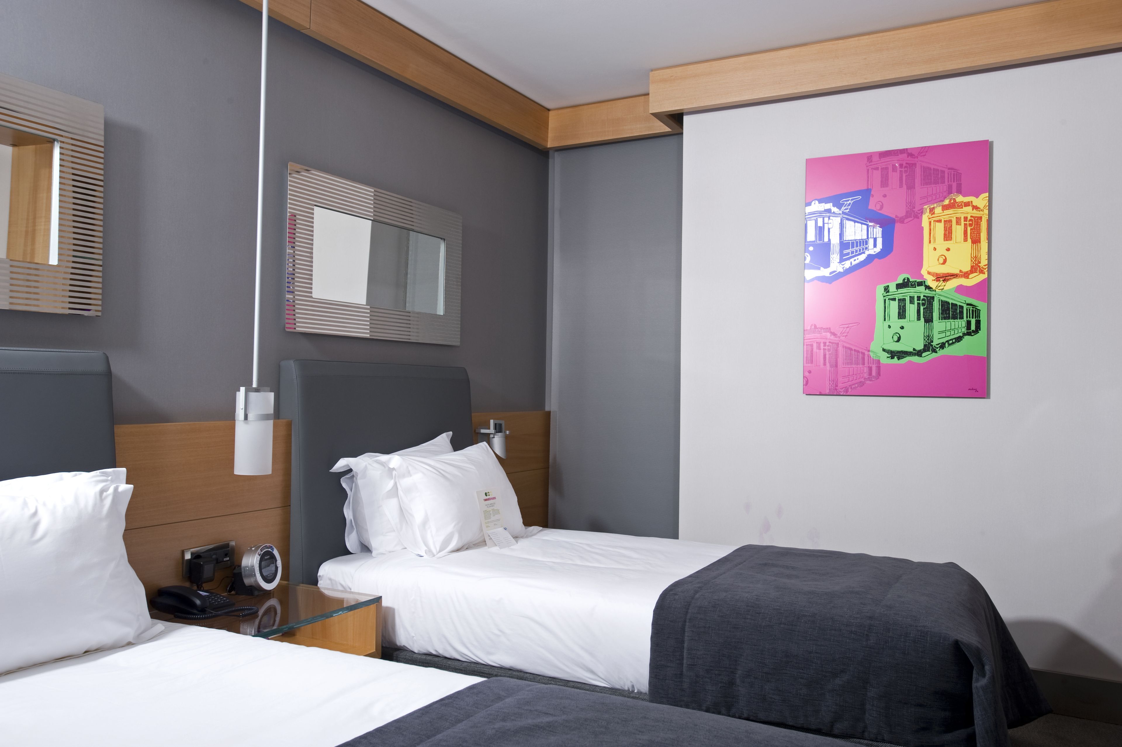 A hotel room with two beds, a bedside table with a phone, a mirror, and a colorful artwork on the wall.