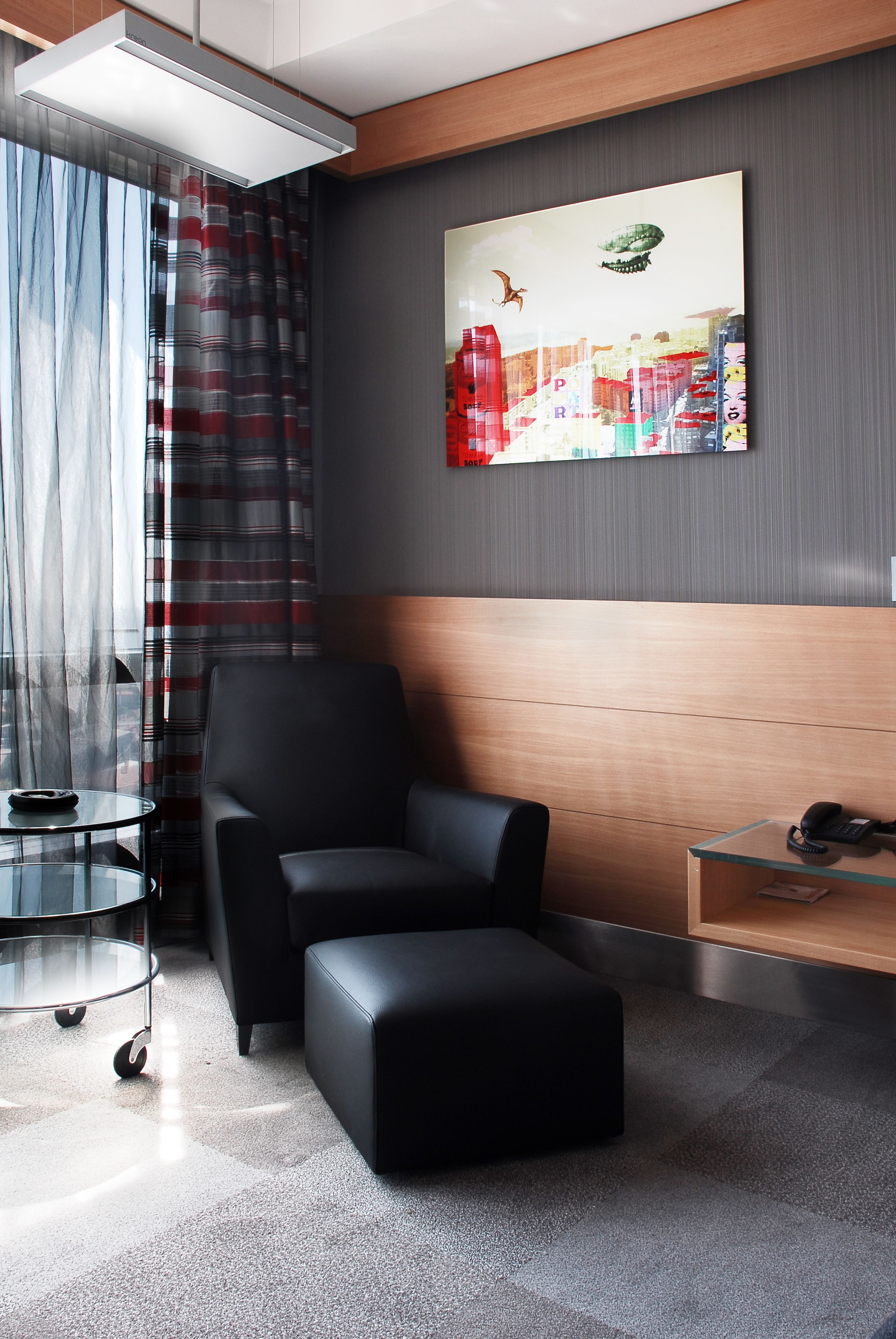 A modern hotel room with a black leather chair, a glass table, and a wooden panel wall.