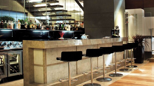 Een moderne bar met een centraal gelegen toonbank, meerdere barkrukken en een trap op de achtergrond.