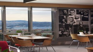 A modern dining area with large windows, a city view, and a wall adorned with black and white photographs. - Point Hotel Barbaros (Istanbul)