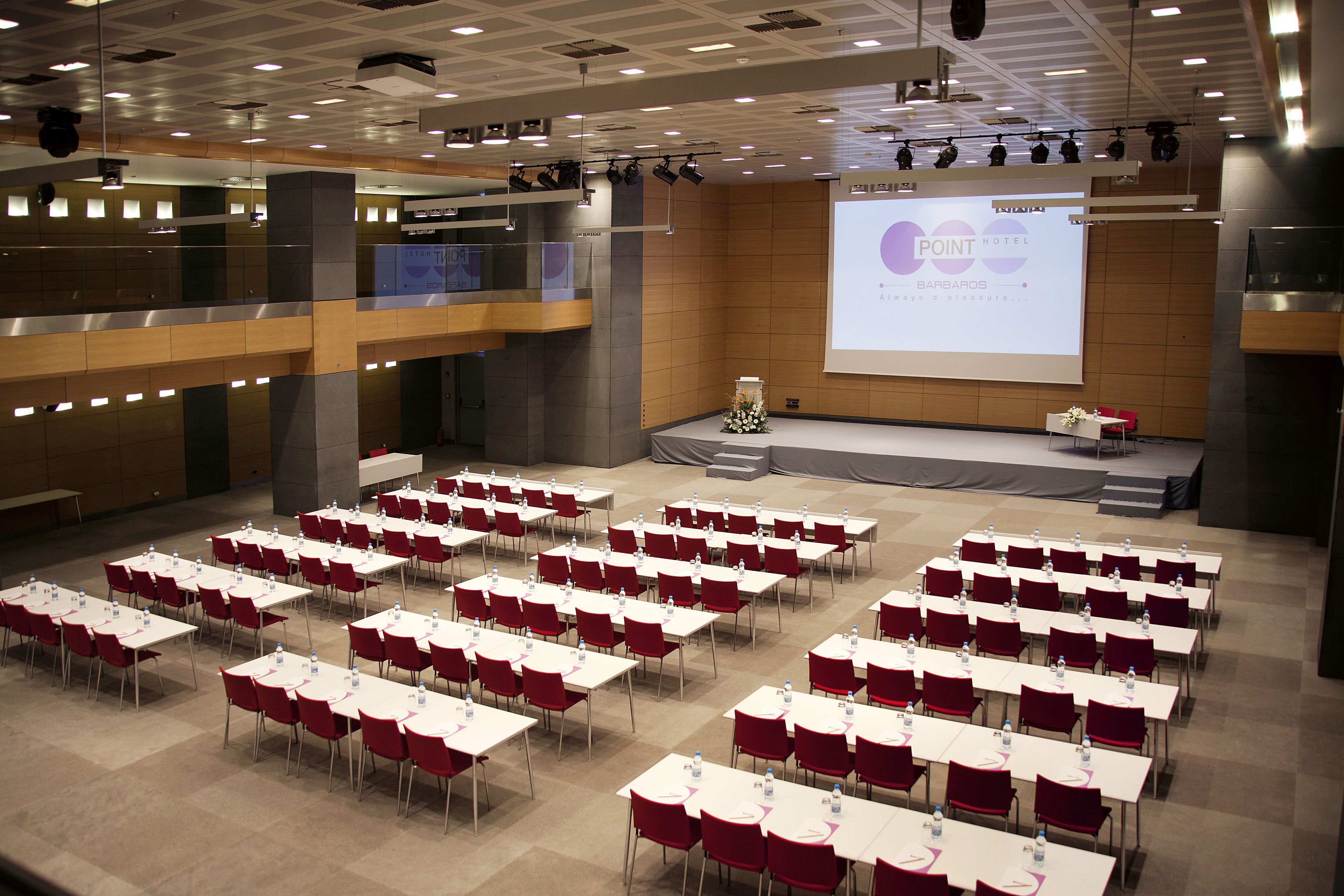 Une salle de conférence dotée de plusieurs tables et chaises, d’une scène avec un écran de présentation et d’un balcon donnant sur la salle.