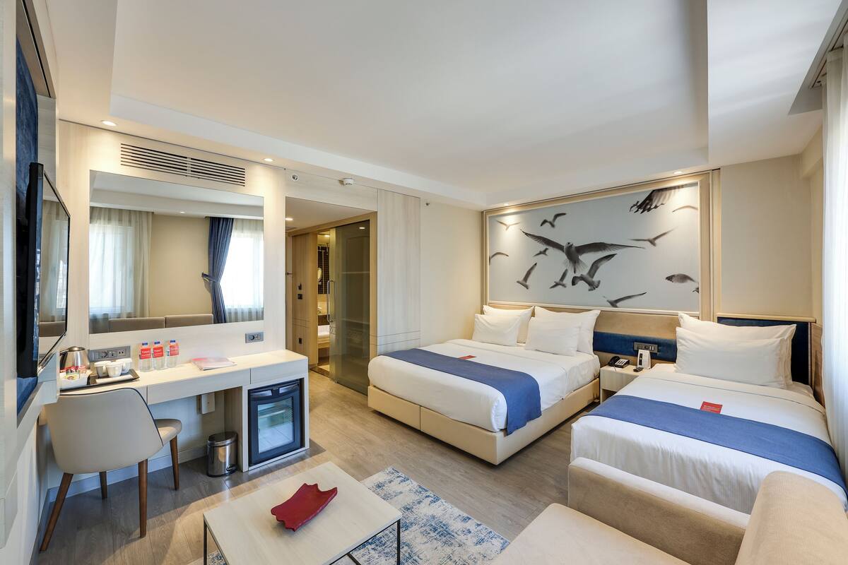 A hotel room with a bed, a desk, a chair, a small fridge, and a painting on the wall.