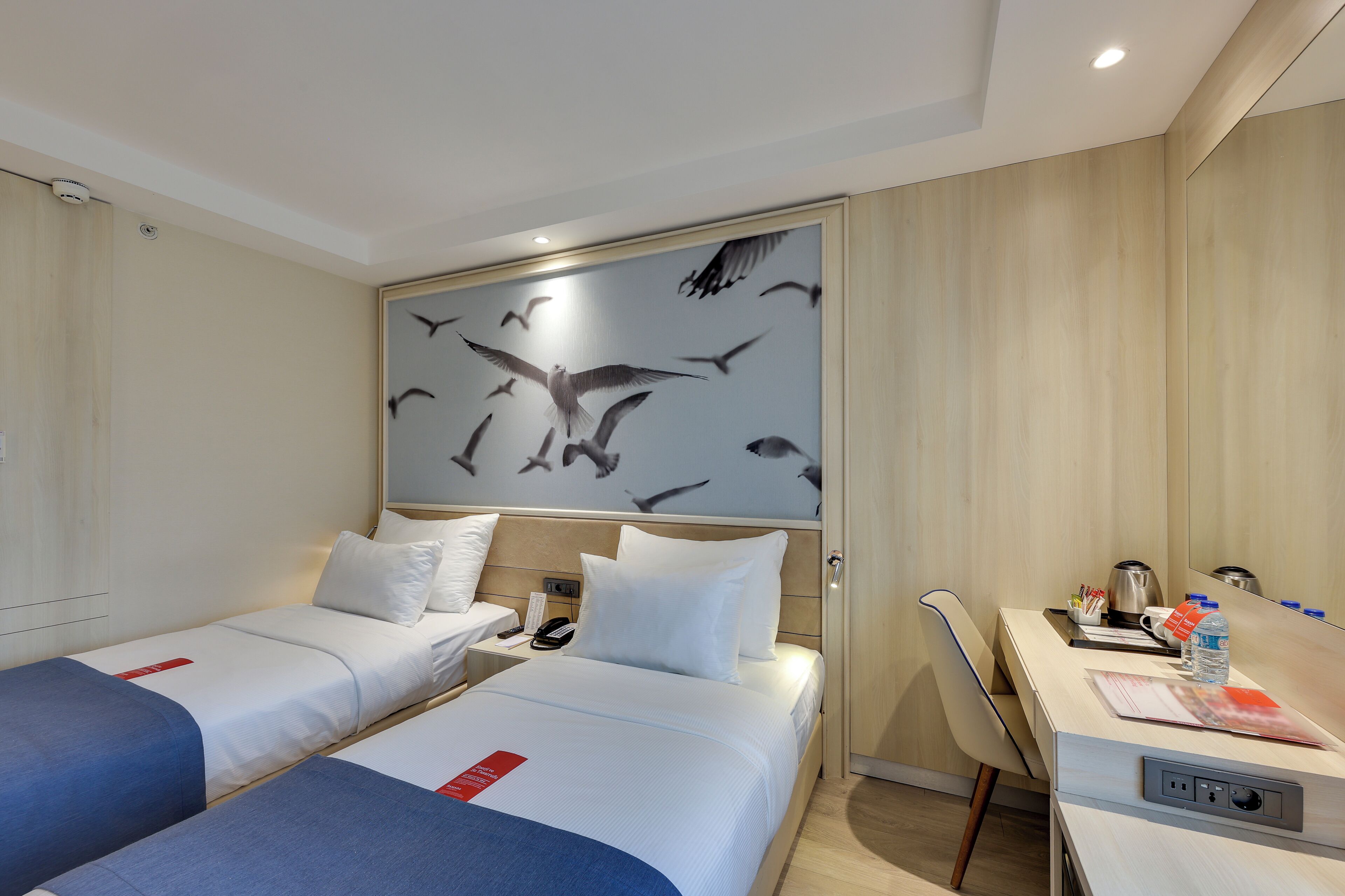 Camera d'albergo con due letti singoli, una scrivania con una sedia, uno specchio e un affresco con uccelli.