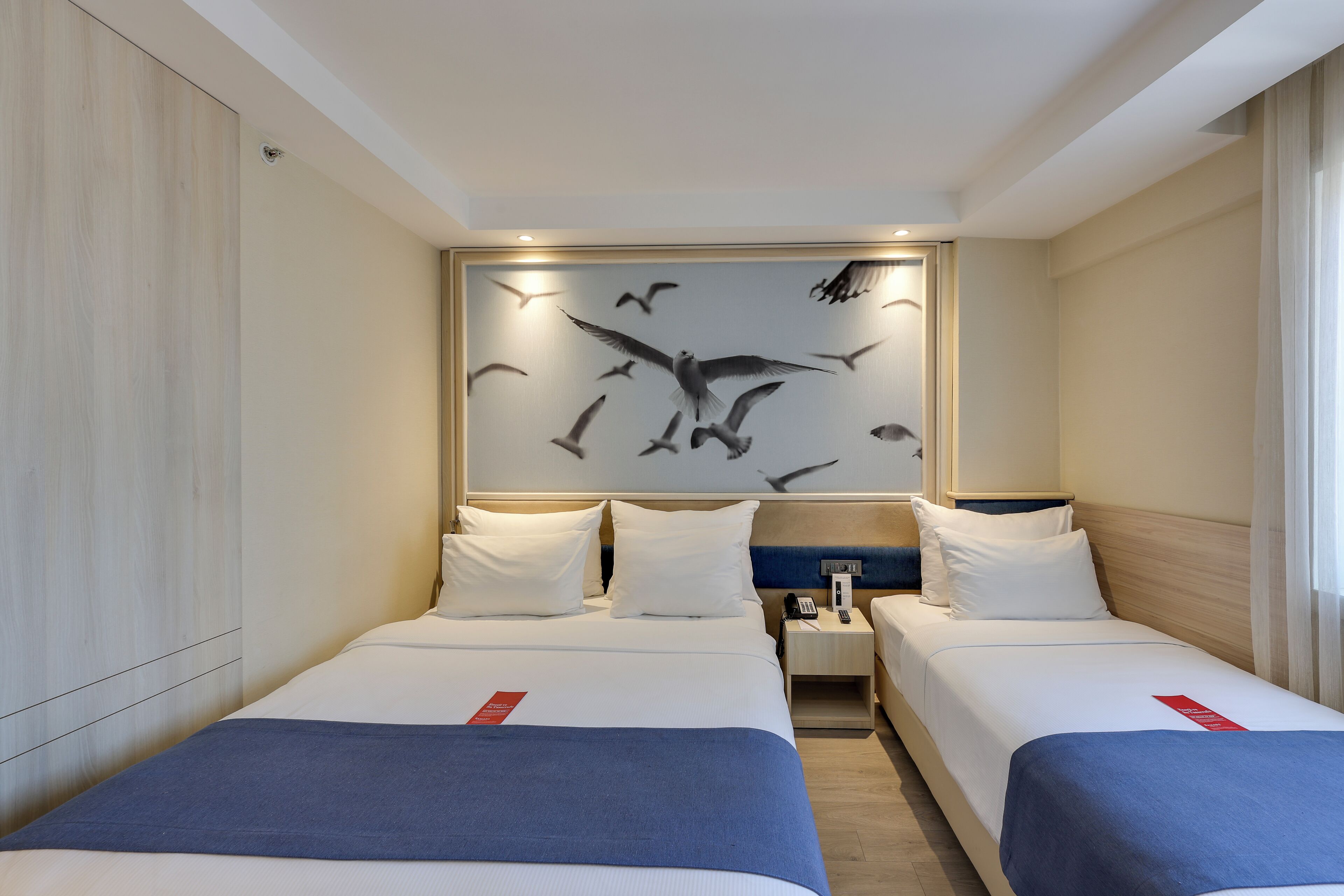 A hotel room with two beds, a wall-mounted artwork of birds, and a nightstand with a phone.