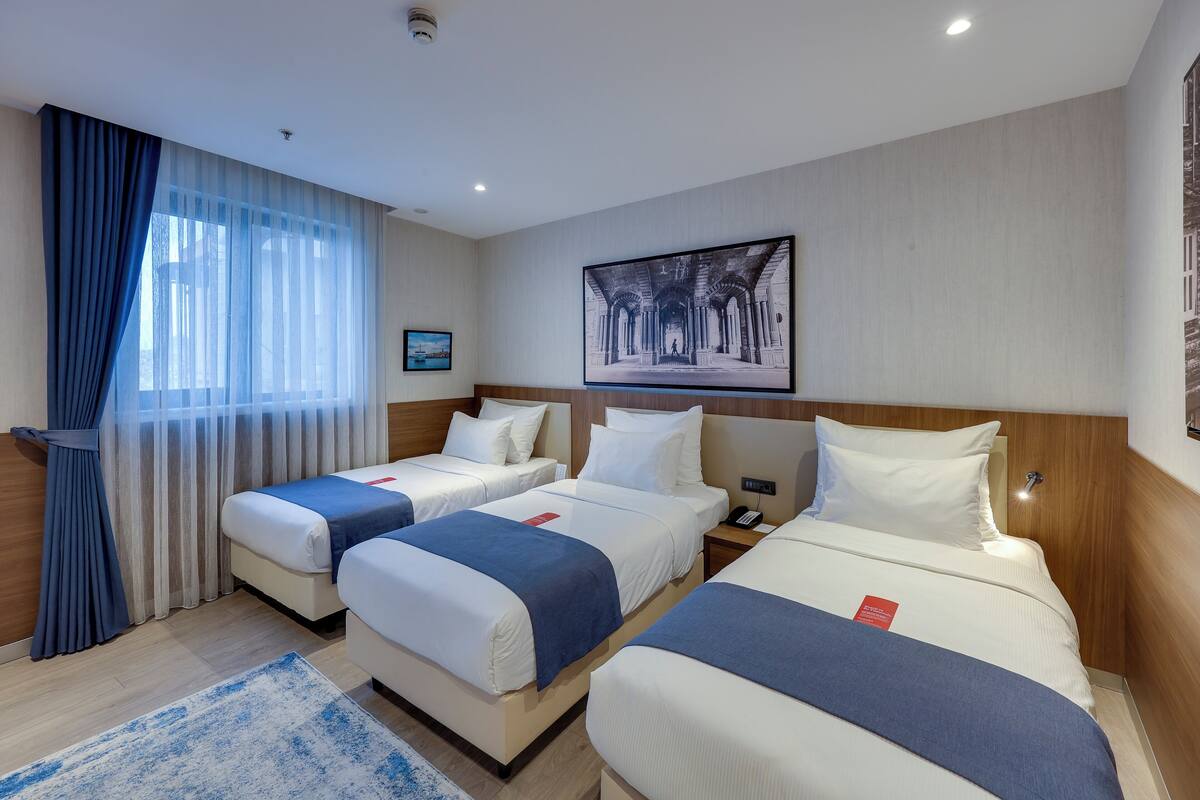 A hotel room with three beds, a large window with sheer curtains, a framed picture on the wall, and a blue rug on the floor.