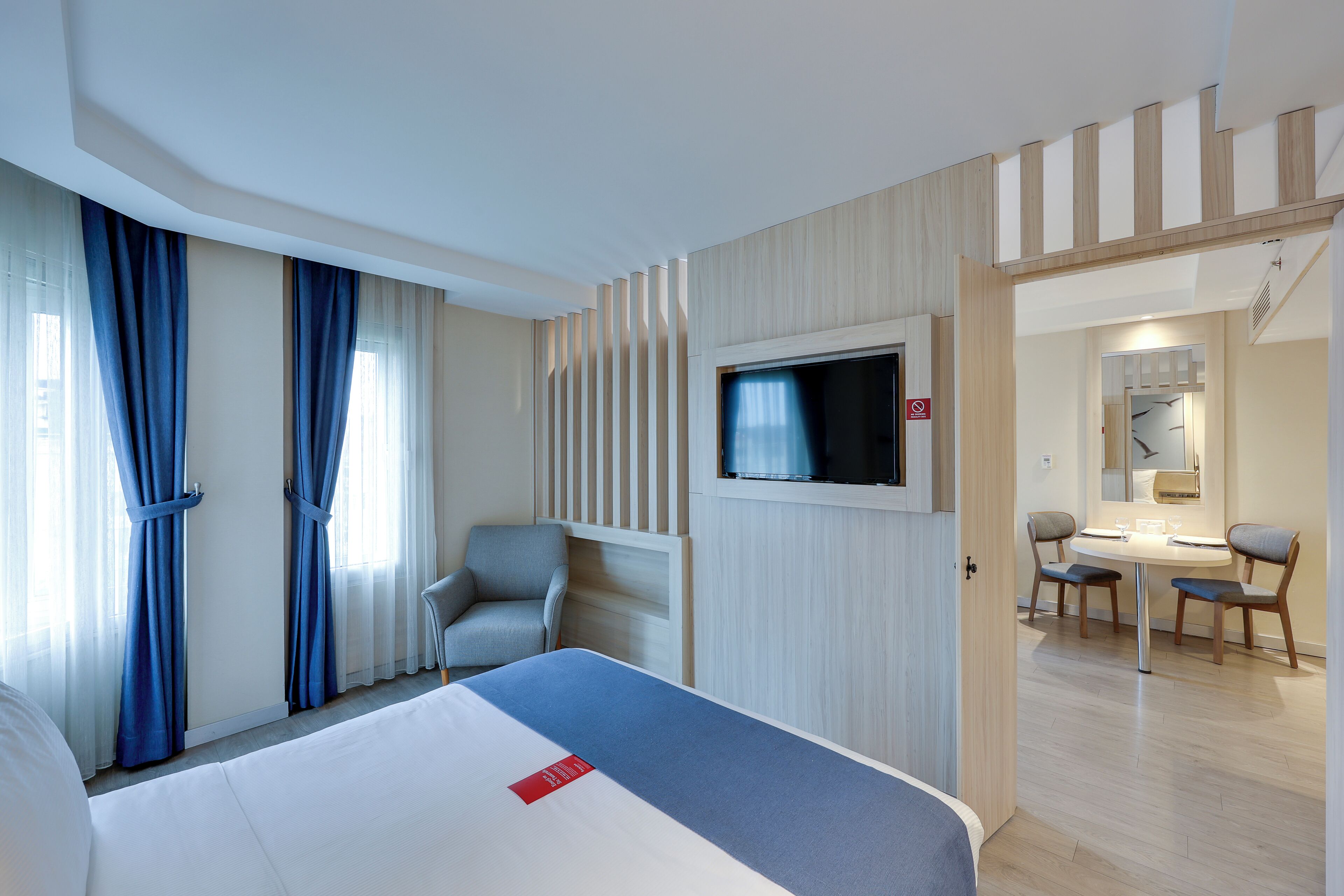 A hotel room with a bed, a TV mounted on the wall, a desk with chairs, and a view of the dining area.