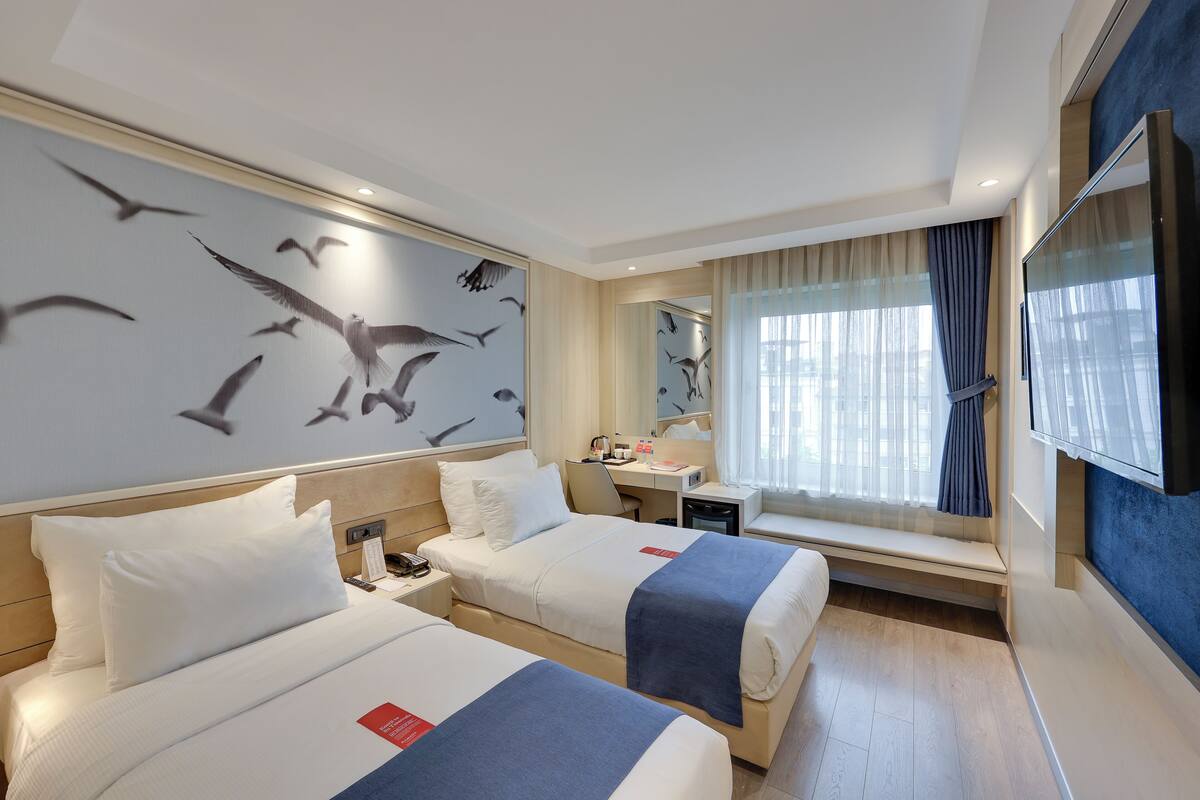 A hotel room with two beds, a desk, a chair, a window with curtains, and a wall with a bird mural.