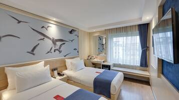 A hotel room with two beds, a desk, a chair, a window with curtains, and a wall with a bird mural.