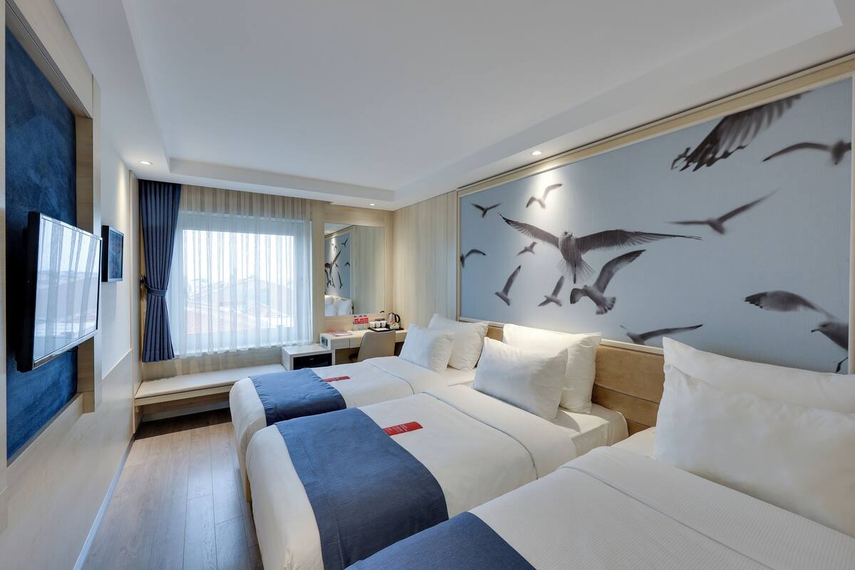A hotel room with two beds, a desk, a chair, a TV, and a large wall mural of birds.