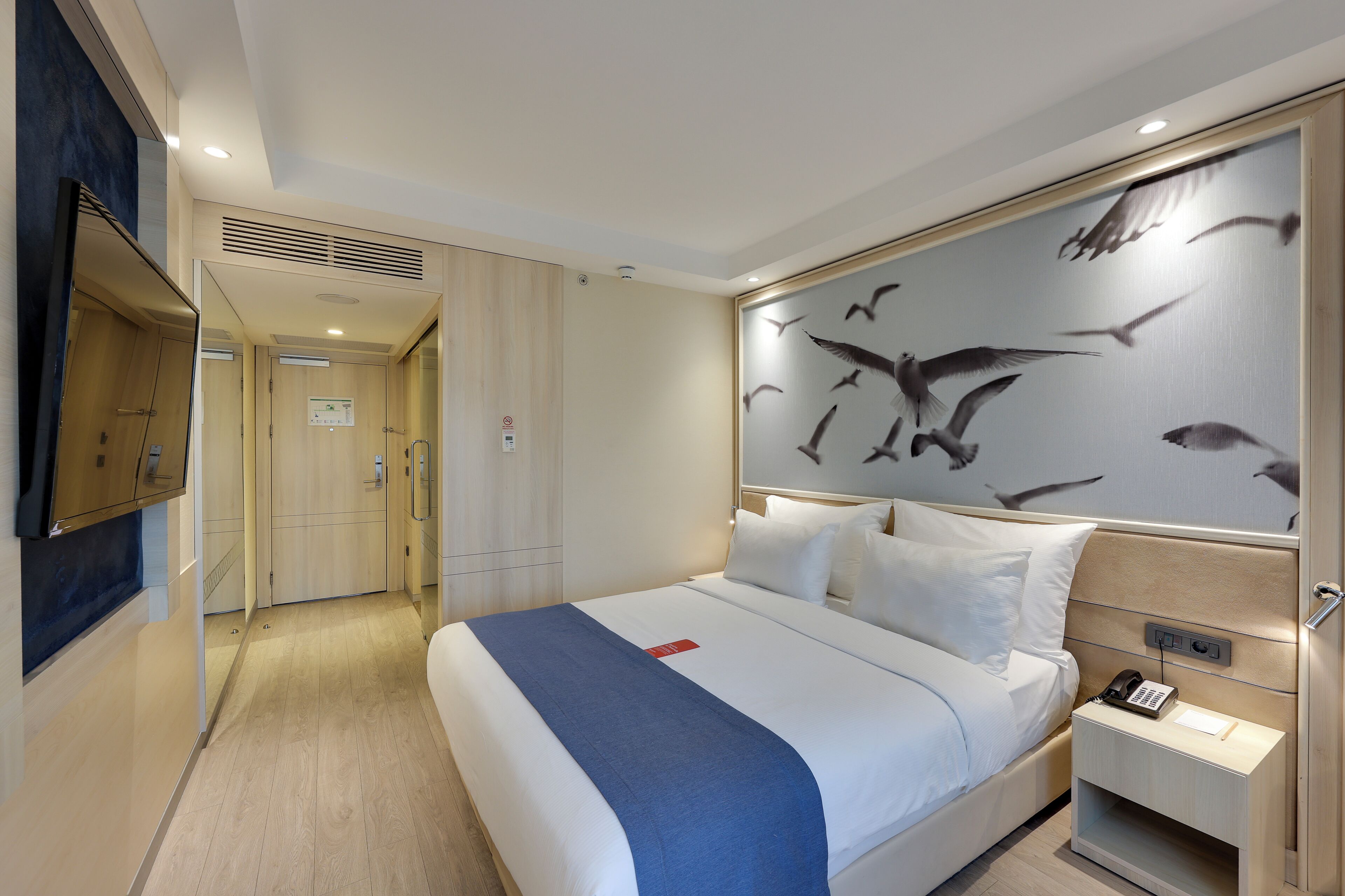 A hotel room with a large bed, a bedside table with a phone, and a wall mural of birds.