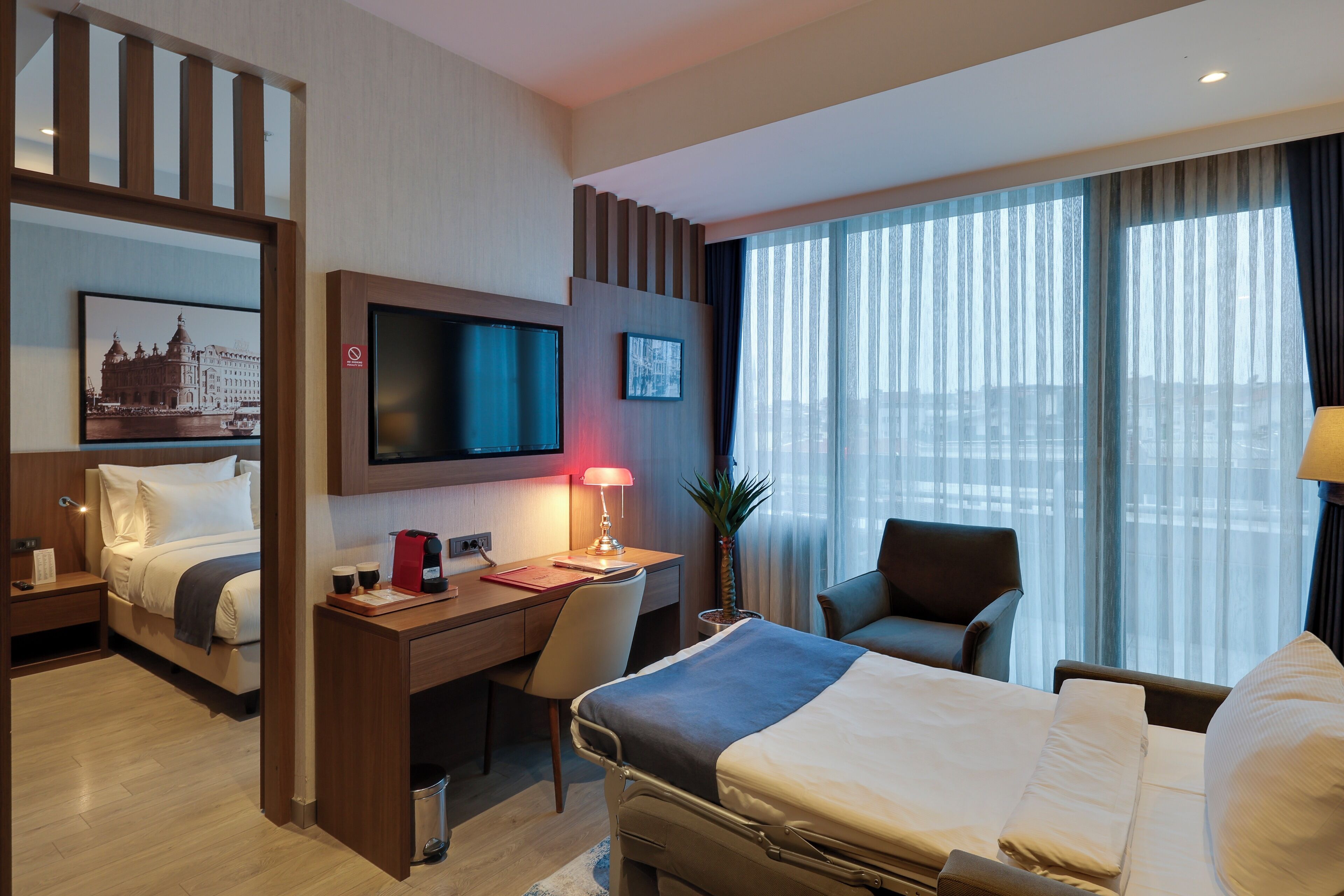 A hotel room with a bed, desk, chair, TV, and large windows.