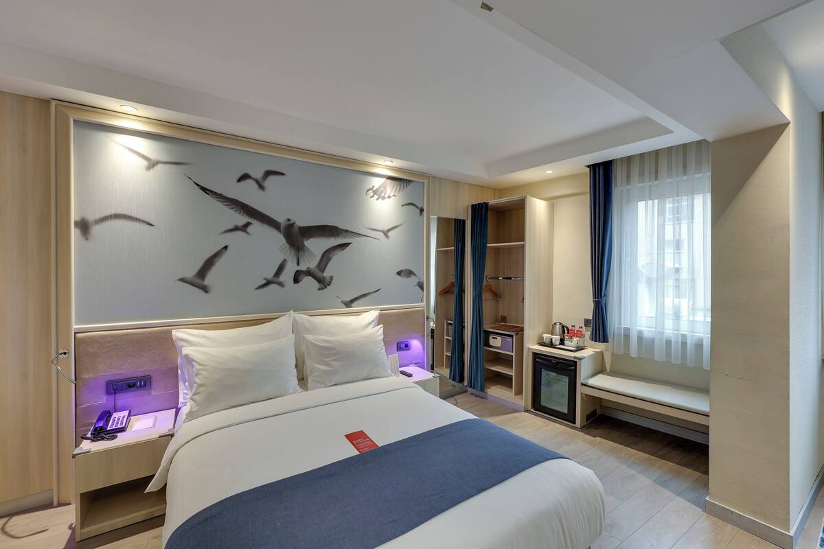 A modern hotel room with a large bed, a built-in desk with a phone, a seating area with a view, and a wall with a bird mural.