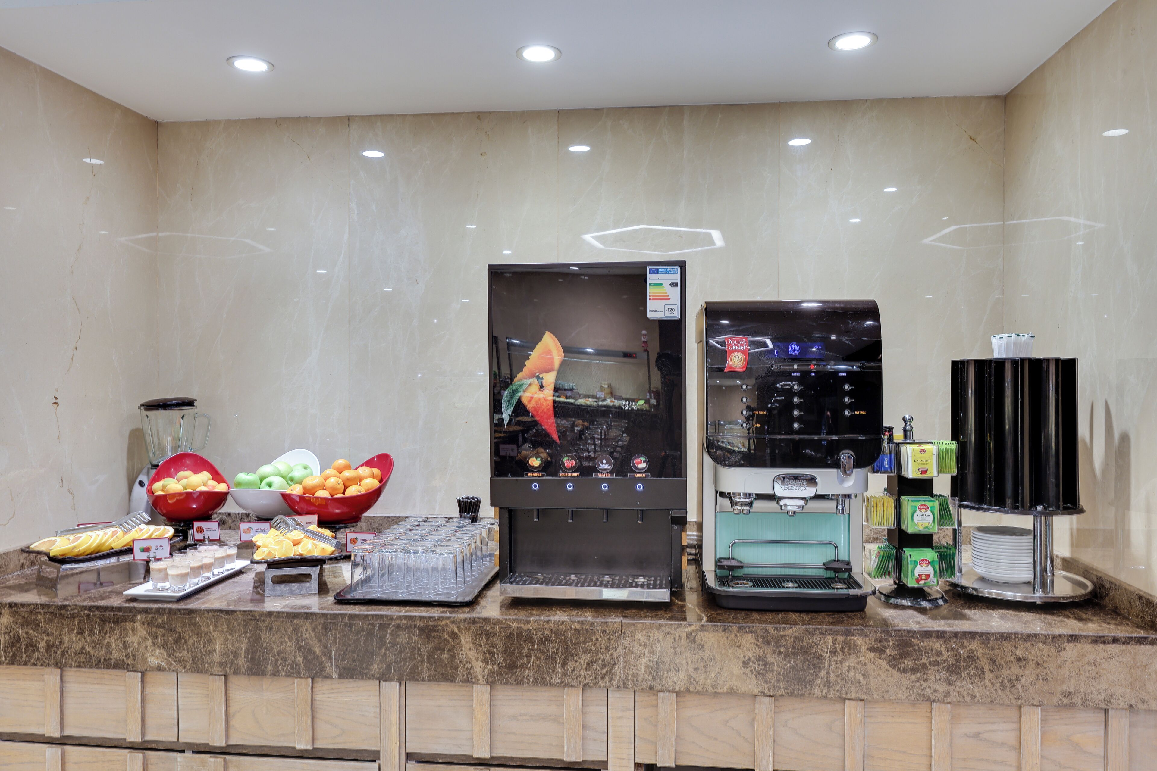 A self-service beverage station with a coffee machine, a juice dispenser, and a refrigerated display of fruits and beverages.
