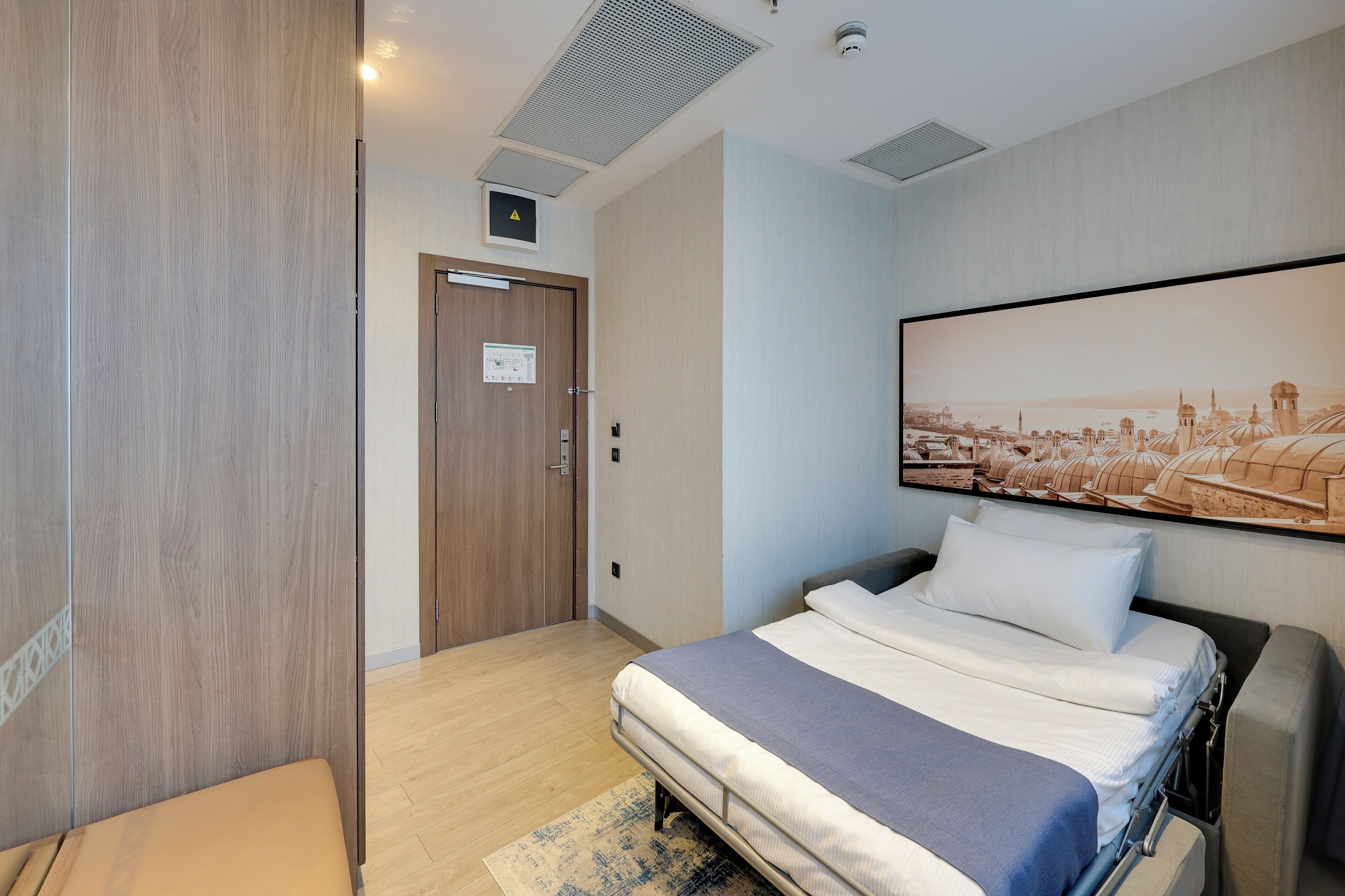 A hotel room with a bed, a wooden wardrobe, a door with a sign, and a cityscape painting on the wall.