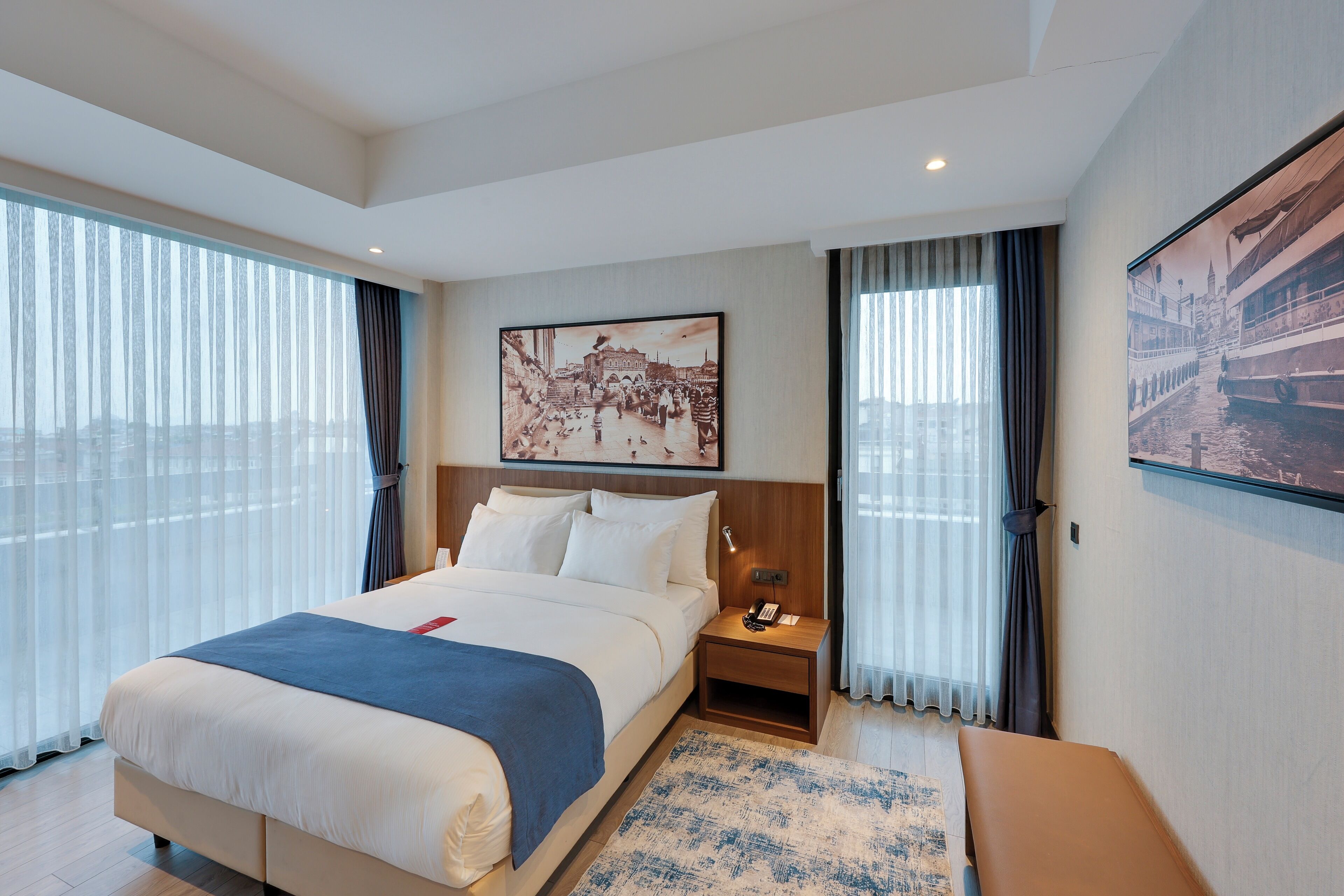 A hotel room with a large bed, a bedside table with a lamp, a nightstand, a window with sheer curtains, and a cityscape painting on the wall.