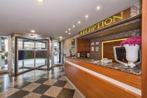 Un área de recepción con piso a cuadros, un mostrador de recepción, una puerta giratoria de vidrio y un letrero que dice "RECEPTION".