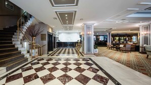 Un vestíbul d'hotel amb un paviment de marbre a quadradets, una escala amb un dibuix de zebra, una recepció i una zona de bar.