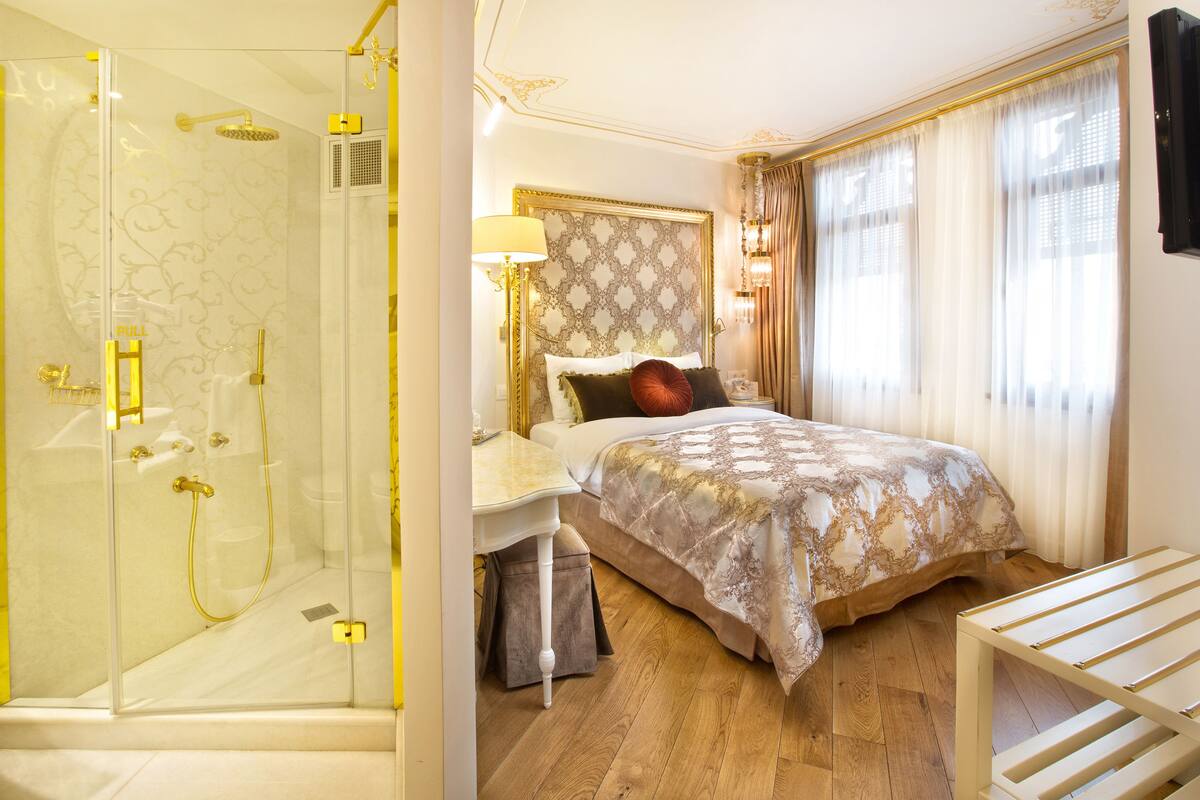 A hotel room with a glass-enclosed shower, a bed with a patterned headboard, and a bedside table with a lamp.