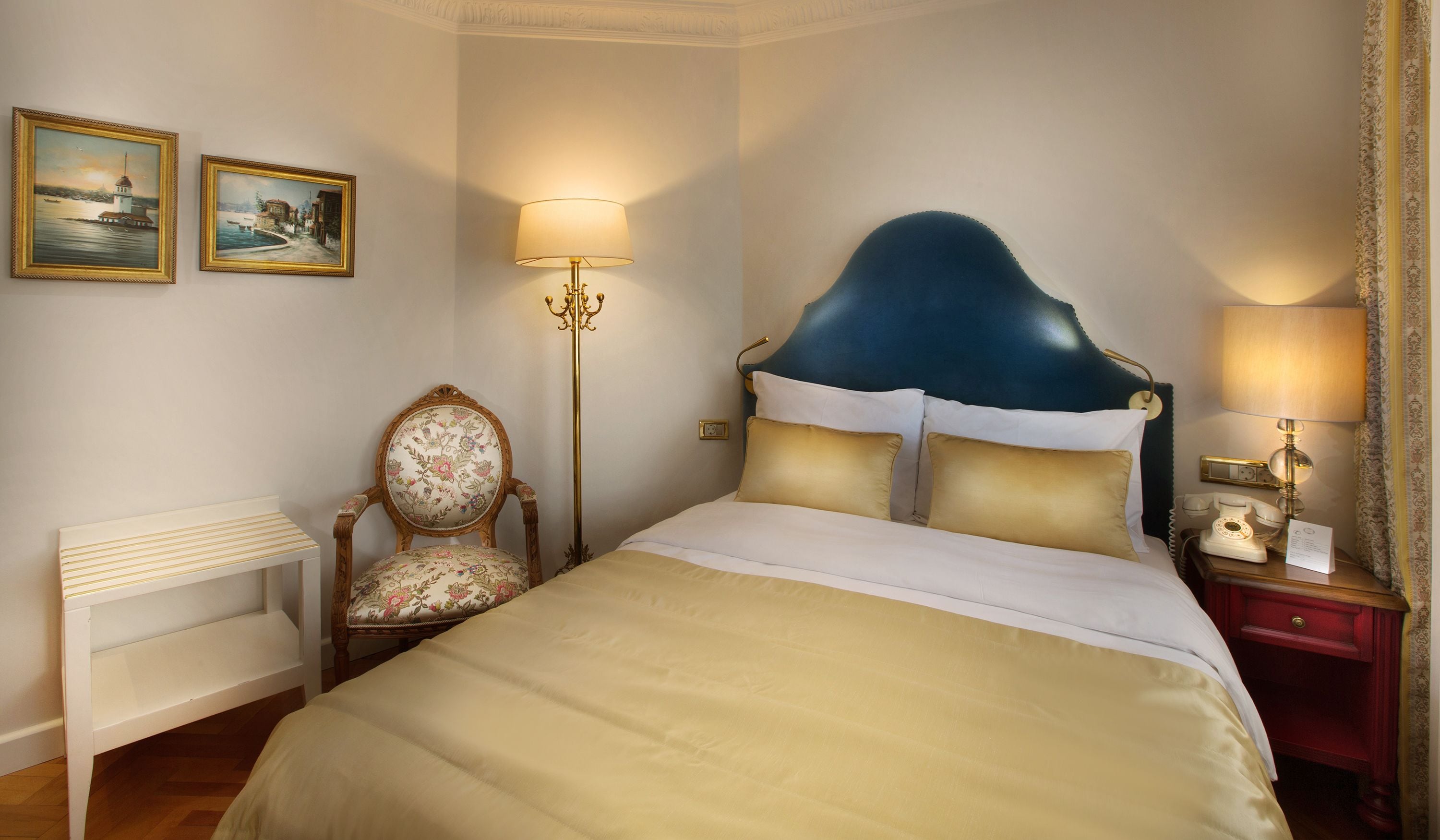A hotel room with a large bed, a chair, two paintings, a lamp, and a nightstand with a telephone.