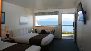 A hotel room with two beds, a desk, a chair, a TV, and a window with a view of the sea. - The Ocean Shores (Homer)