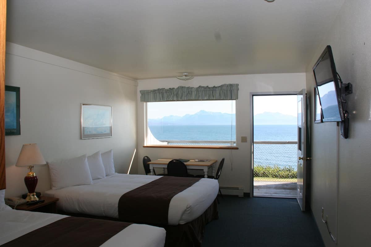 A hotel room with two beds, a desk, a chair, a TV, and a window with a view of the sea.