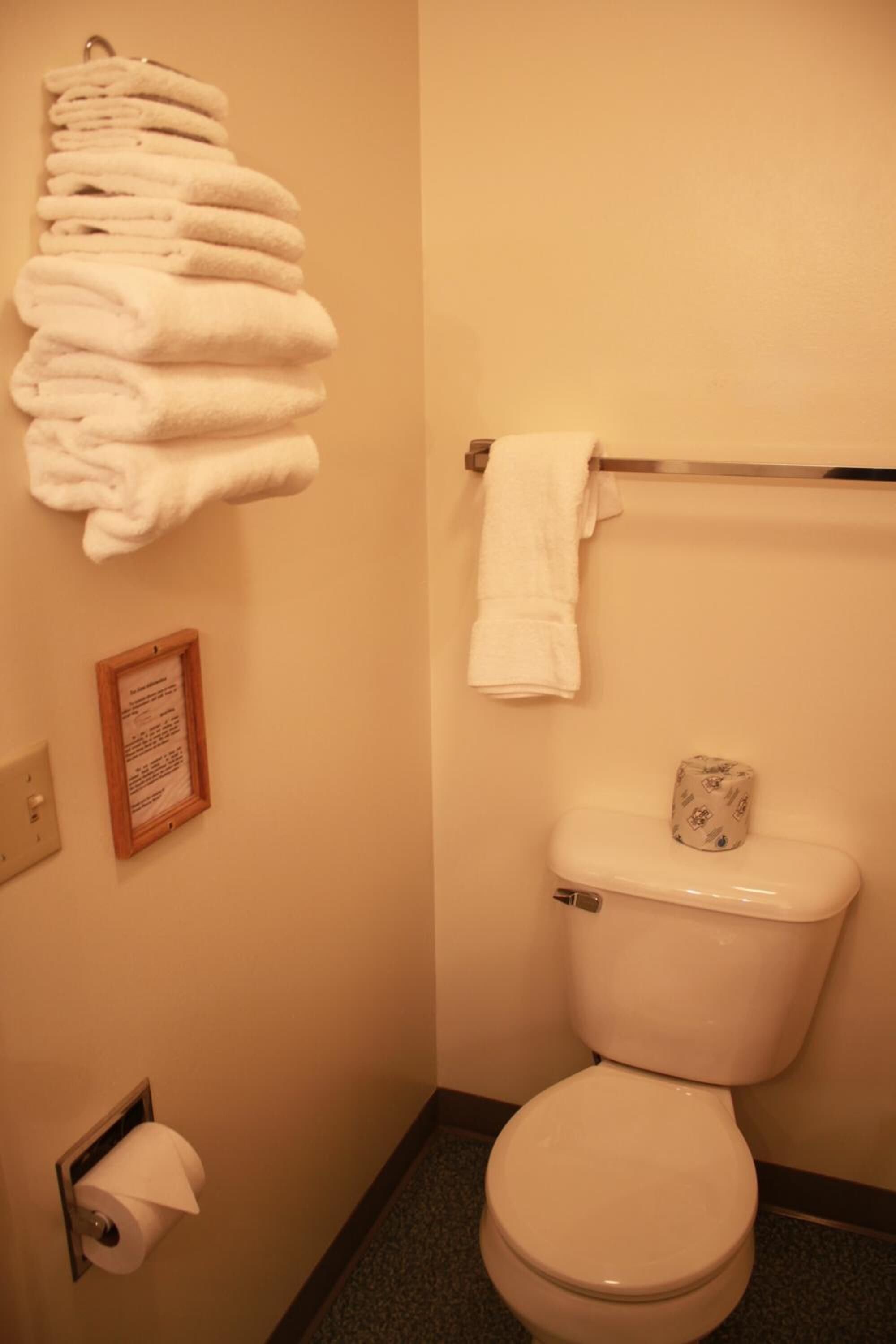 A bathroom with a toilet, a stack of white towels, a framed sign, and a toilet paper holder.
