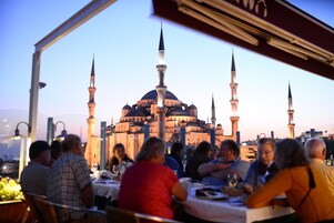 A restaurant with tables set for dining, a view of a mosque with multiple minarets, and a cityscape in the background.