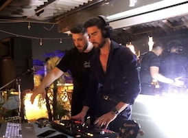 Dos personas operando equipo de DJ en una habitación tenuemente iluminada, con palmeras visibles y un techo rústico.