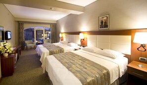 A hotel room with two beds, a desk with a lamp, a TV, and a window with curtains.