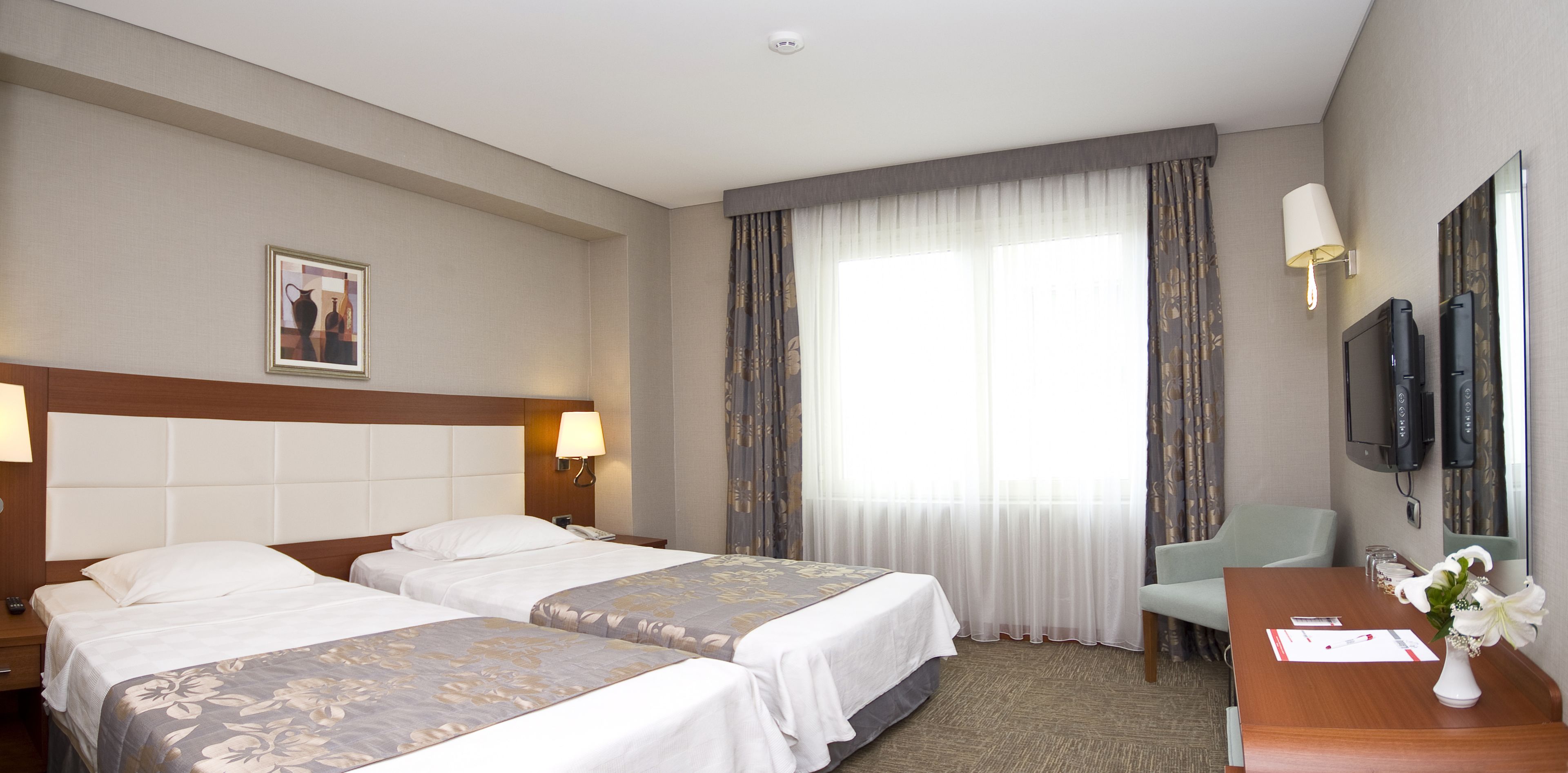 A hotel room with two beds, a desk with a vase of flowers, a television, and a large window with curtains.