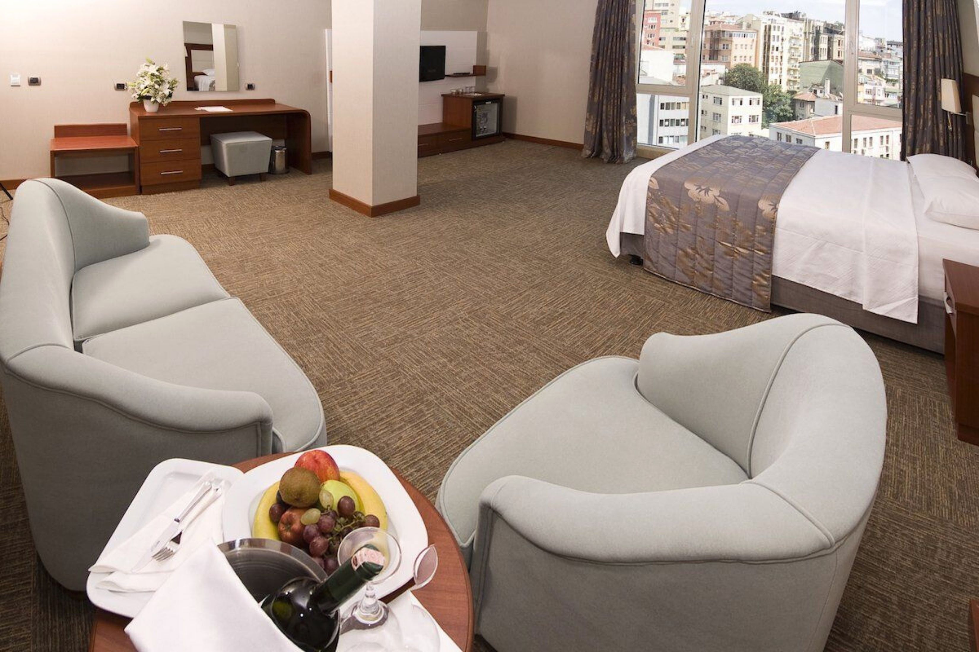 A hotel room with a bed, two armchairs, a desk with a mirror, a small TV, and a view of the city.