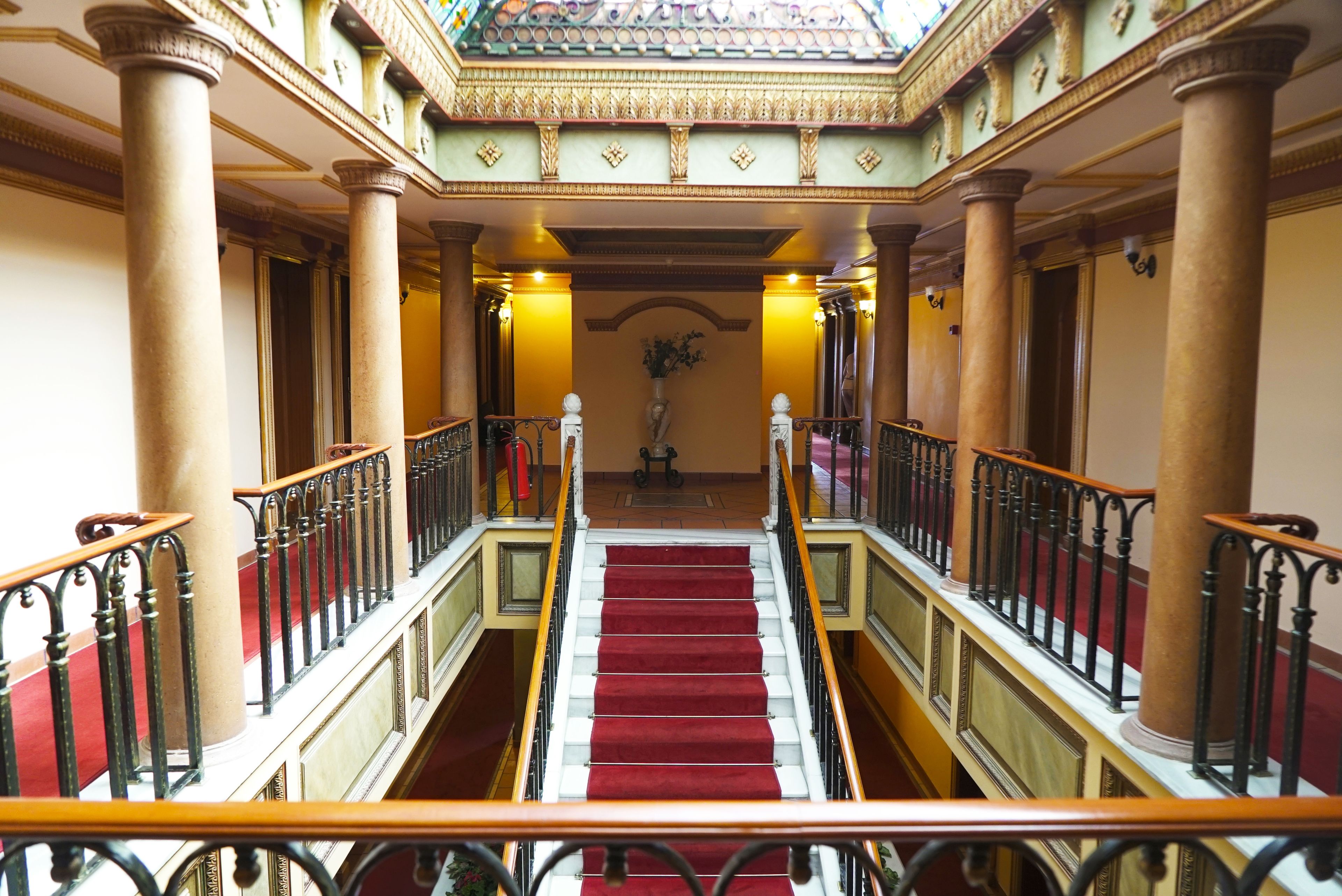 Een statige trap met rood tapijt, omzoomd door zuilen en versierde balustrades, die leidt naar een ruimte met hoge plafonds, een decoratieve boog en een bloevas.