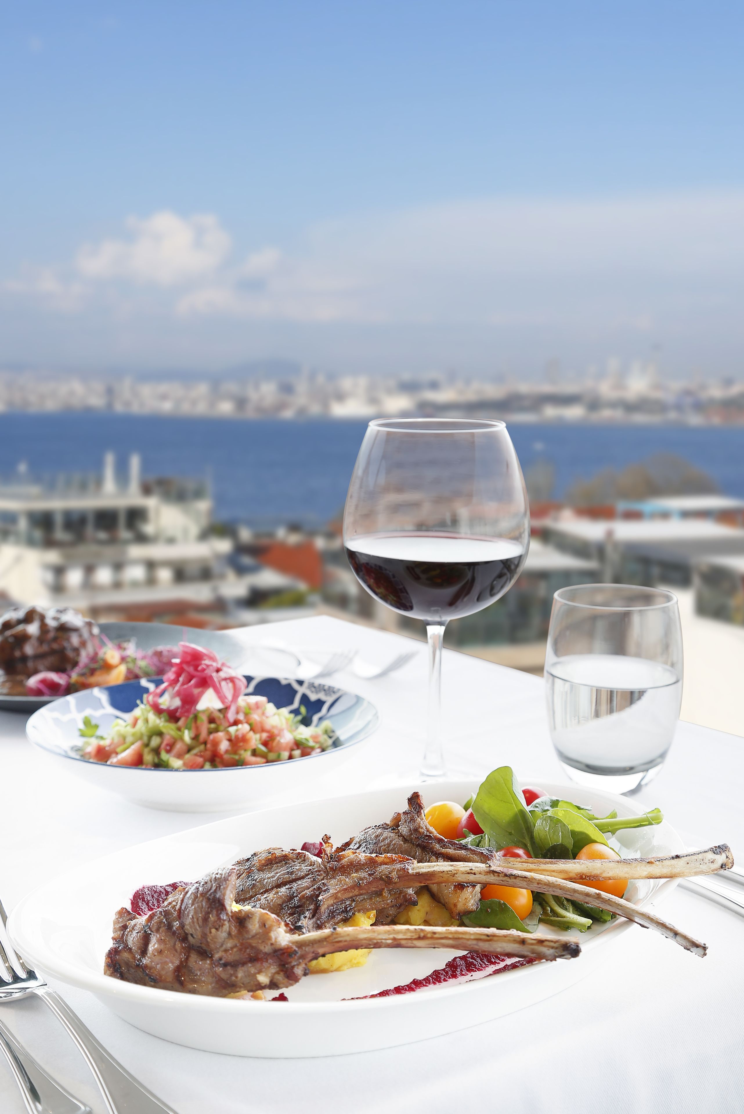 Una mesa con un plato de chuletas de cordero, ensalada y una copa de vino tinto, frente a una vista panorámica de la ciudad.