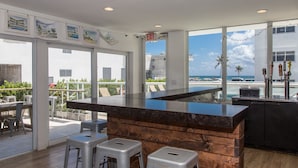 A modern bar area with a wooden counter, two bar stools, and large windows offering views of the ocean and buildings. - Premiere Hotel (Fort Lauderdale)