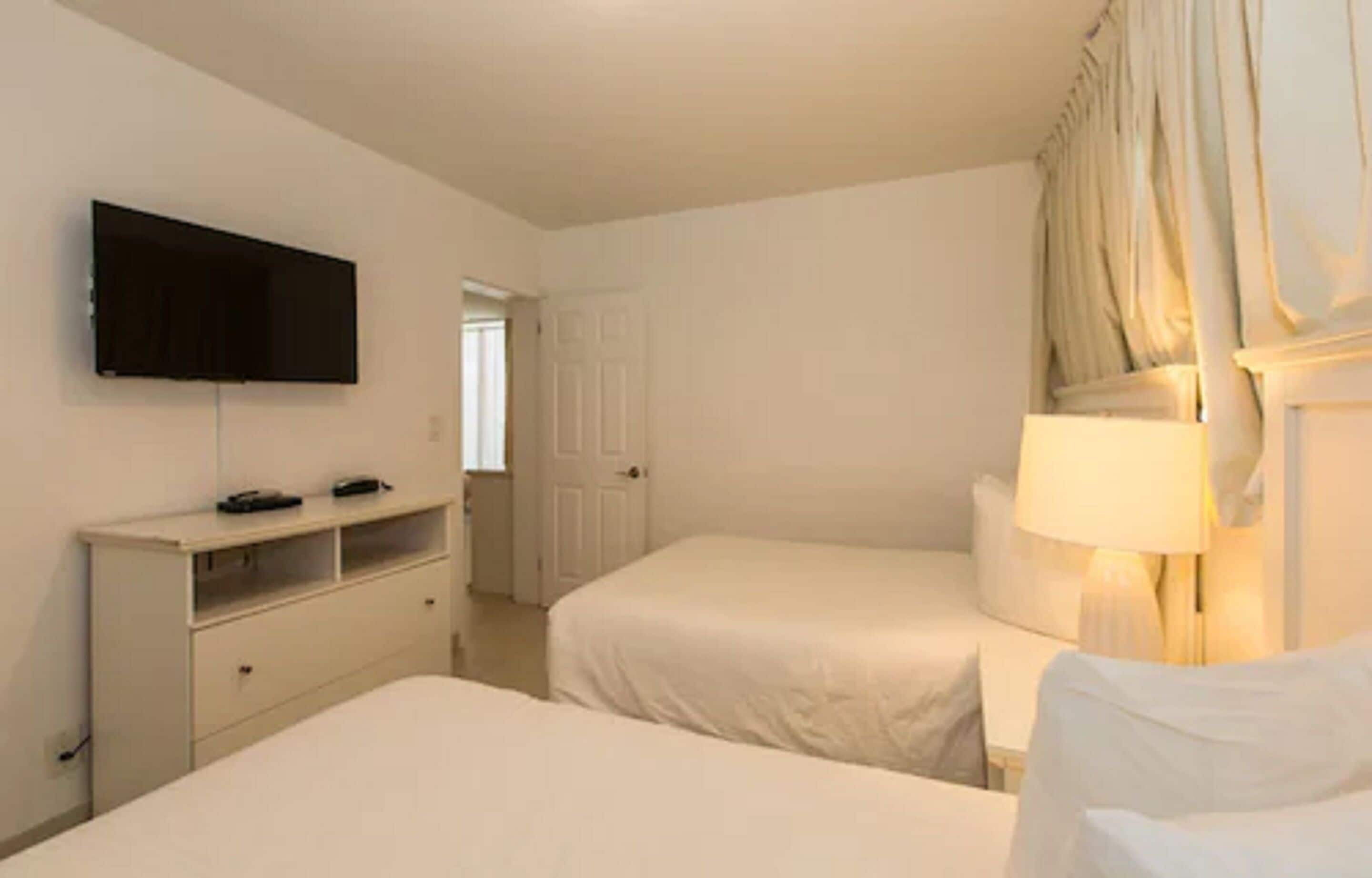 Een hotelkamer met twee bedden, een aan de muur bevestigde televisie, een nachtkastje met een lamp en een kledingkast.
