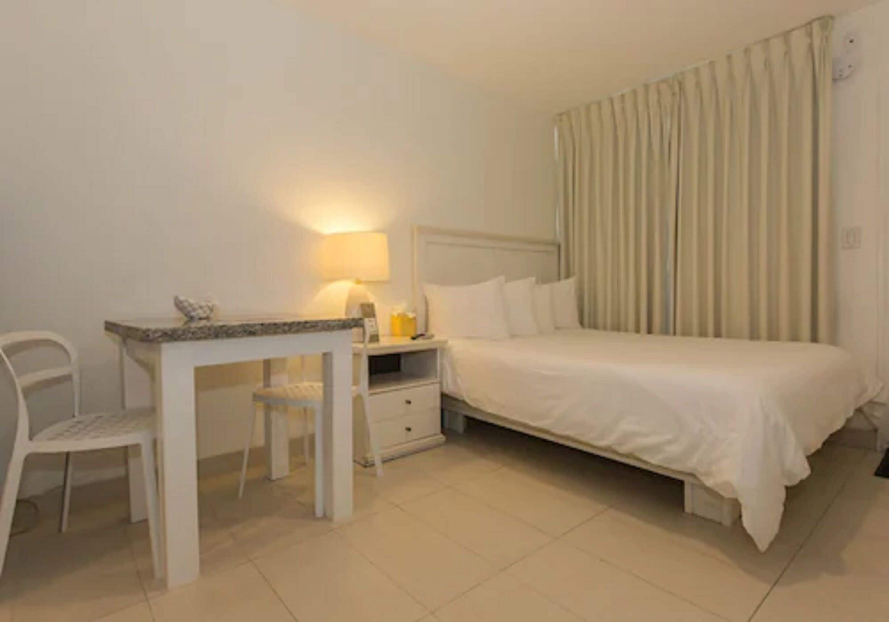 Una camera d'albergo con un letto, una scrivania con una sedia, una lampada e un comodino.