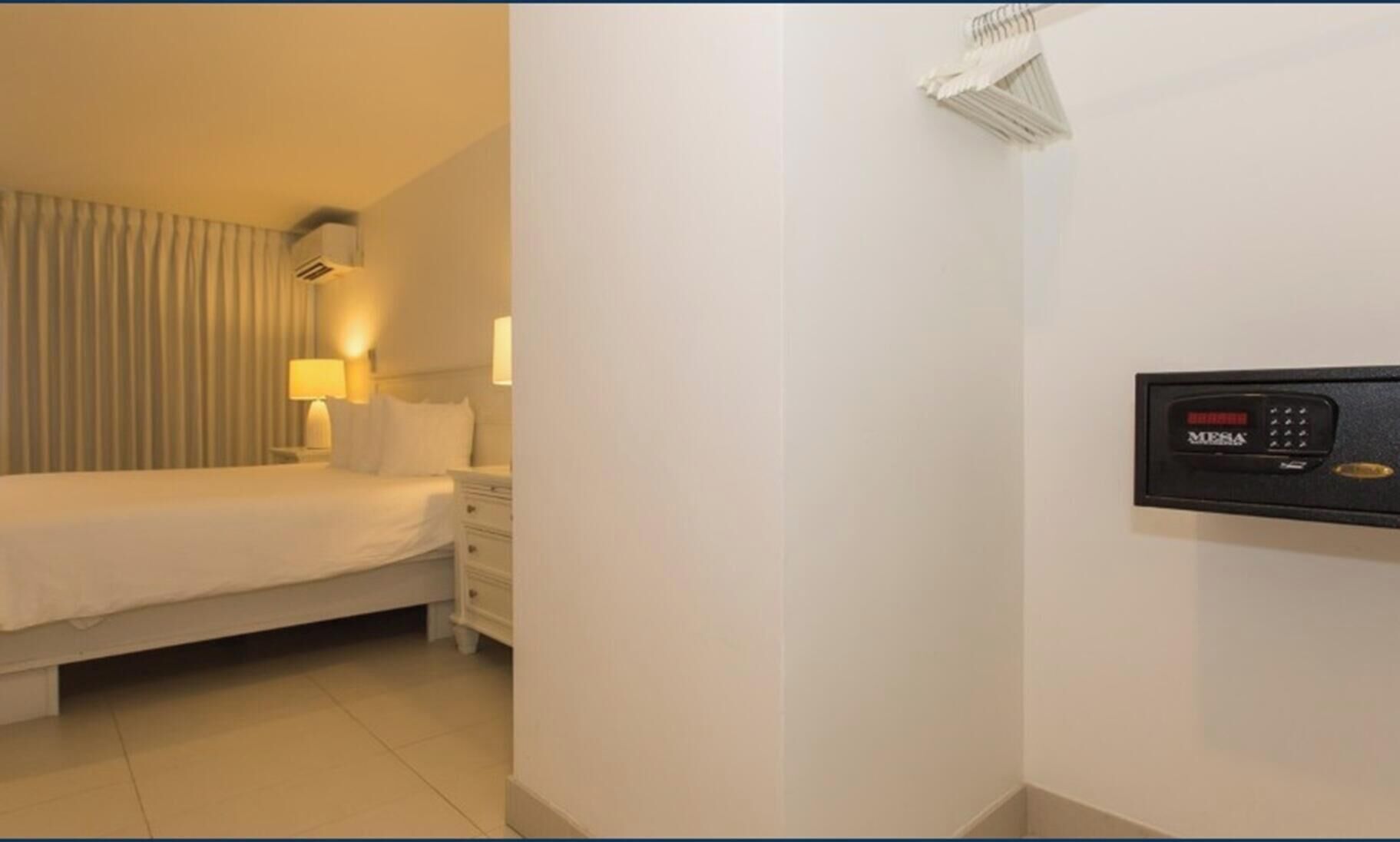 Hotellihuone, jossa on sänky, yöpöytä, lamppu ja seinässä turvapaneeli.