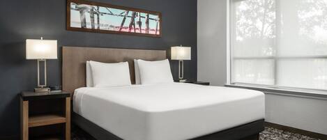 Une chambre d’hôtel bien rangée, avec un grand lit, deux tables de chevet avec des lampes, un tableau encadré au mur et une fenêtre avec des stores.