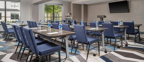 A hotel conference room with tables, chairs, water bottles, and a TV on the wall.
