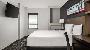 A hotel room with a bed, a desk, a chair, a TV mounted on the wall, and a window.