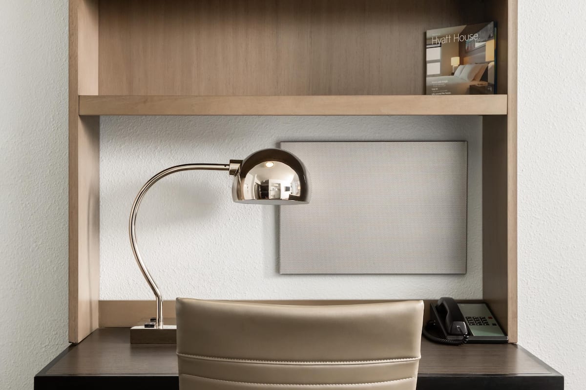 Ett hotellrum med ett skrivbord, en lampa, en telefon, en bok med titeln ”Hyatt House” och en väggmonterad skärm.