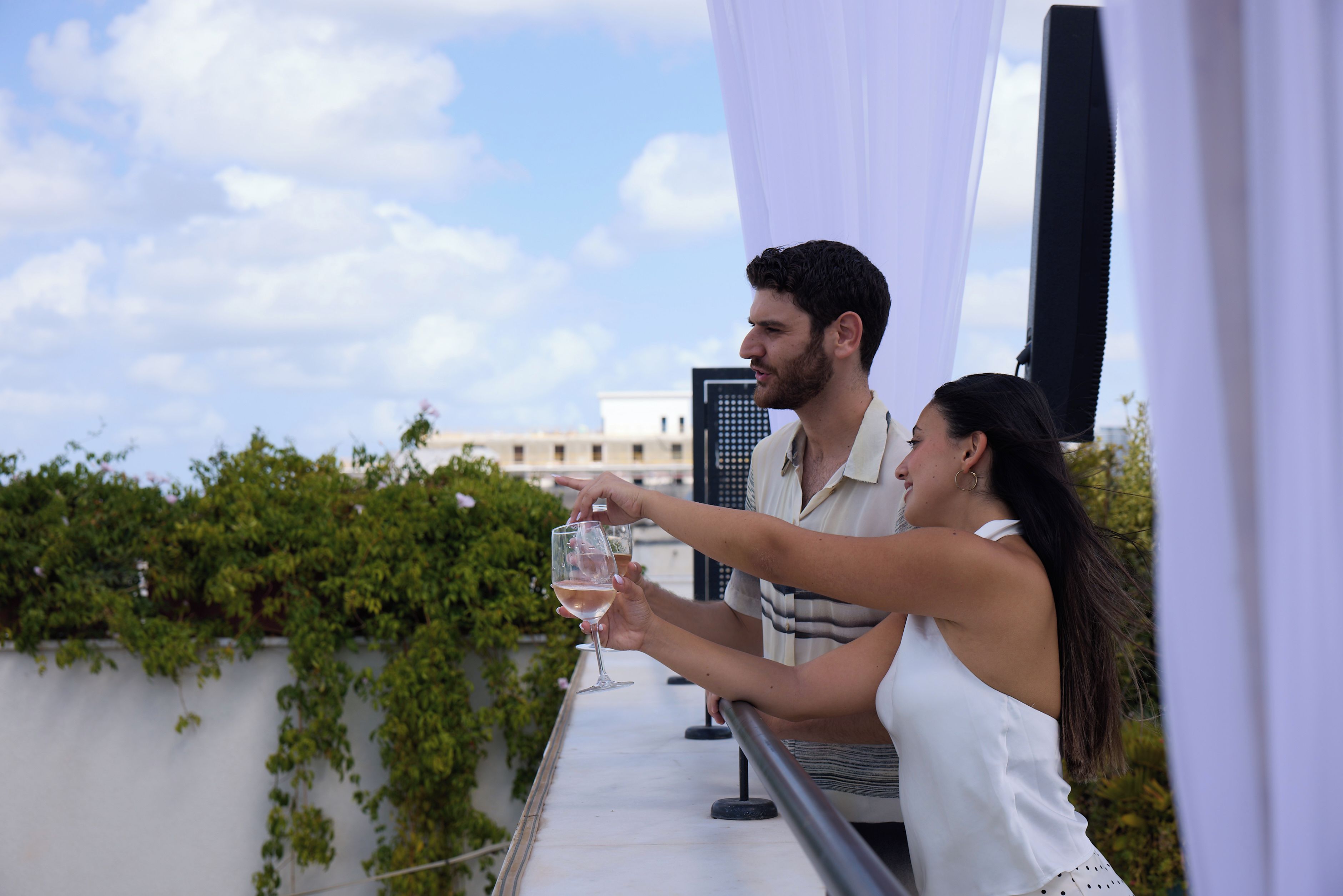 Um casal brindando com taças de vinho em um terraço ao ar livre, com vista para prédios e vegetação.