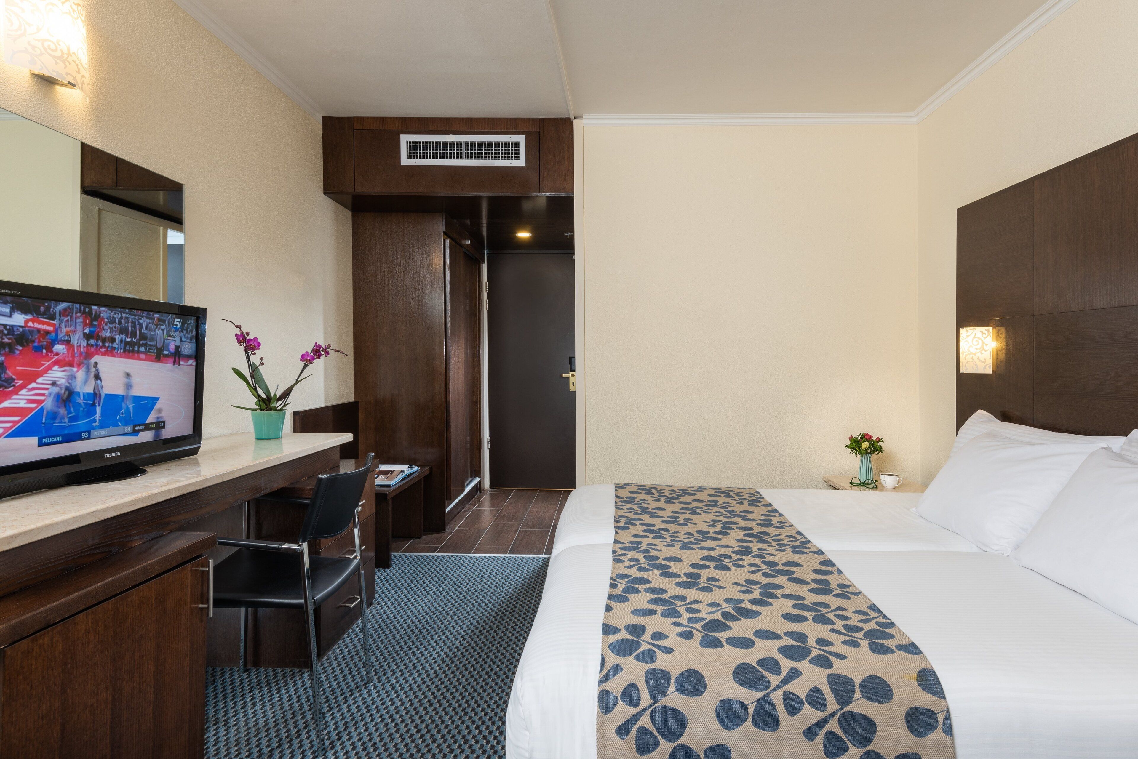 A hotel room with a bed, a desk with a television, a chair, a potted plant, and a nightstand with a lamp.