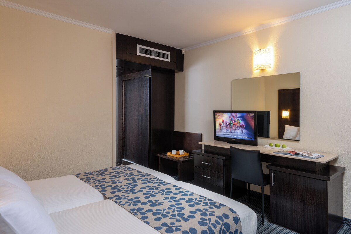 Une chambre d’hôtel avec un lit, un bureau, une télévision, un miroir et une petite table sur laquelle se trouvent une tasse et un livre.