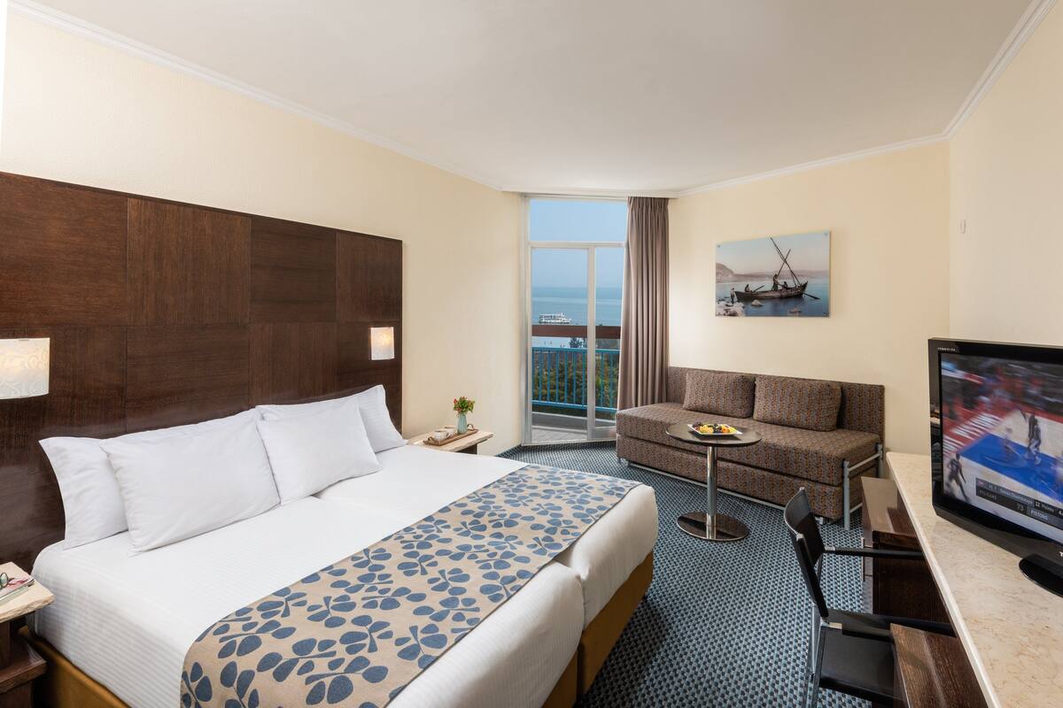 A hotel room with a large bed, a sofa, a small table, a TV, and a balcony with a view of the sea.