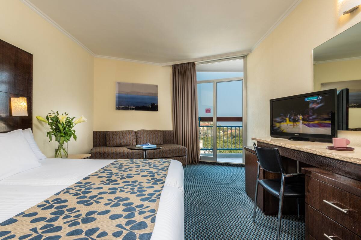 A hotel room with a large bed, a sofa, a desk with a television, and a view of the outdoors.