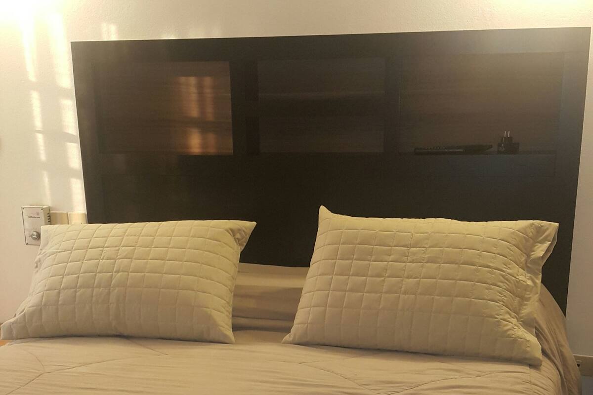 A bed with a blue bedspread and two pillows, a dark headboard, and a wall-mounted light fixture.