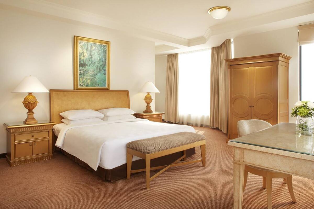 A hotel room with a large bed, two bedside tables with lamps, a wooden bench, a glass table, a wardrobe, and a painting on the wall.