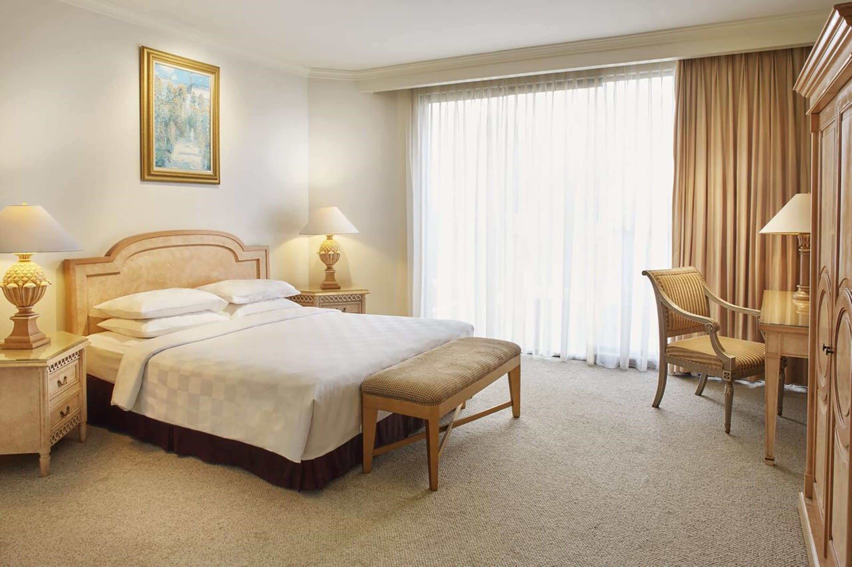 Habitación de hotel con una cama grande, dos mesitas de noche con lámparas, una silla, un banco, un armario y un ventanal con cortinas.