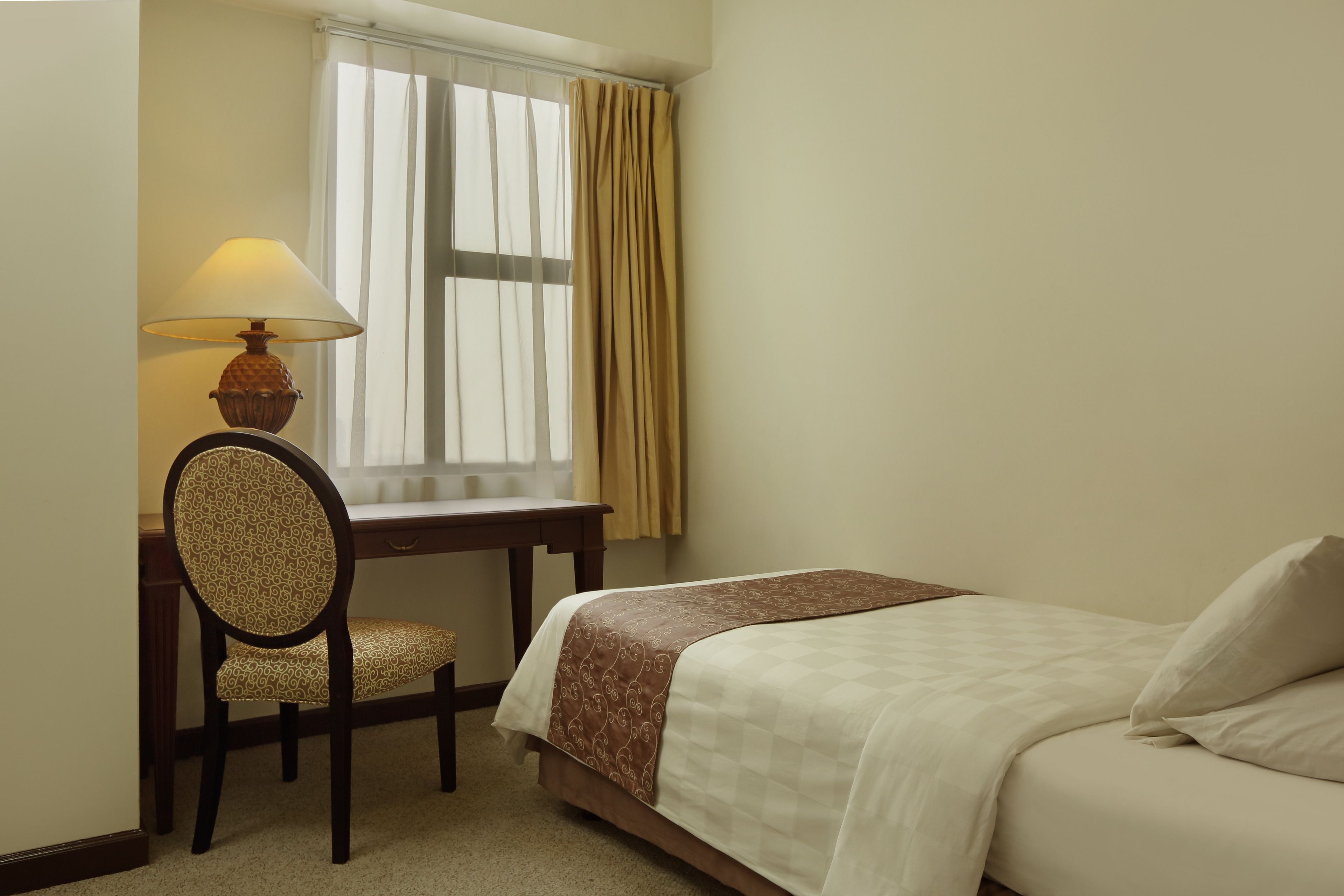 A bedroom with a bed, a desk with a chair, a lamp, and a window with curtains.